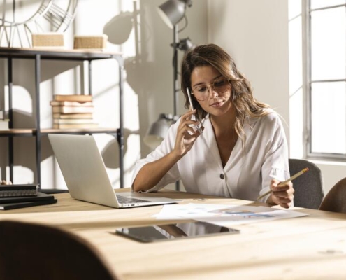 Imagem de uma mulher loira com camiseta branca, senta atras de uma mesa com um notebook e papelada, falando ao celular e anotado coisas no papel