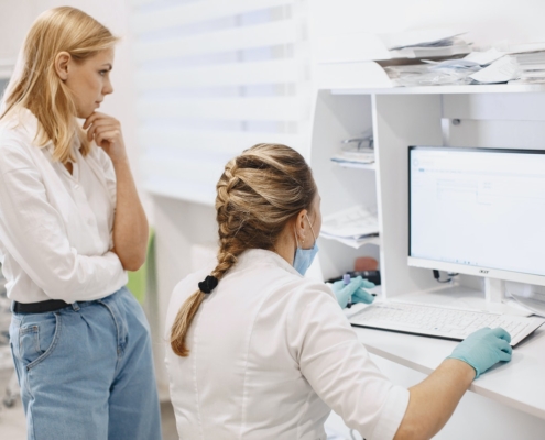 Imagem de duas mulheres olhando na tela de um computador. Um delas está em pé e a outra sentada.