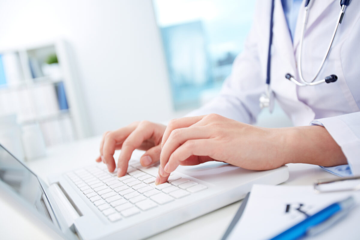 mãos de uma médica vestindo jaleco branco e estetoscópio mexendo em um notbook branco. O balcão em que está apoiado onotebook também é branco e o fundo é um hospital bem ilumiando e desfocado na imagem, porém prevalece a cor branca