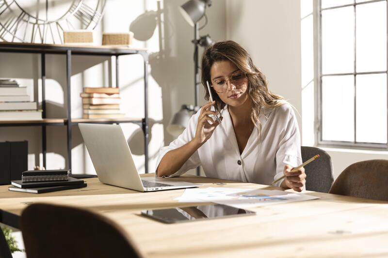 Imagem de uma mulher loira com camiseta branca, senta atras de uma mesa com um notebook e papelada, falando ao celular e anotado coisas no papel