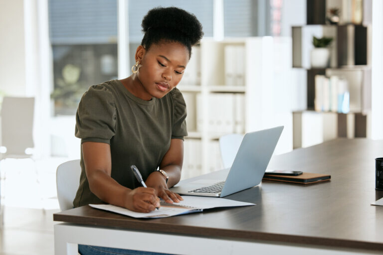 mulher sentada em frente a uma mesa com seu notebook e escrevendo no papel